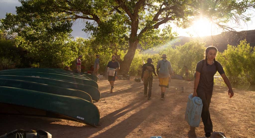 A group of people carry supplies by a row of canoes resting on land. 